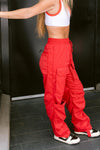 Adult dancer wearing red cargo-style dance pants, standing indoors with one hand on hip.