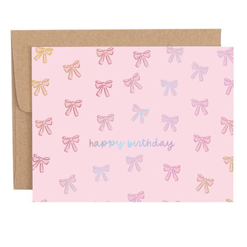 Pink birthday greeting card with bow pattern and holographic foil accents, shown with a kraft envelope behind it