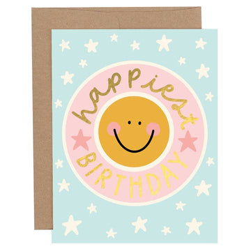Birthday greeting card with a pink smiley face and the words “Happiest Birthday” in gold foil lettering on a light blue background with white star details, shown with a brown envelope.