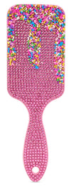 A pink hairbrush covered in rhinestones, with a handle and paddle head decorated with colorful sprinkle-like embellishments on a white background.