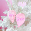 Two pink glitter BFF heart Christmas ornaments hanging on a white holiday tree.