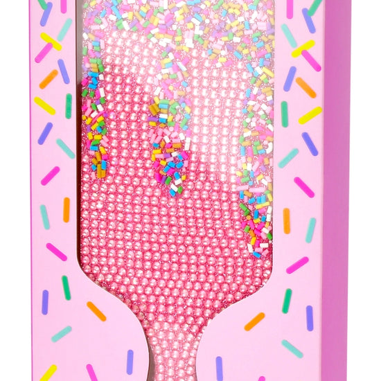 Pink rhinestone-embellished paddle brush in its retail packaging, featuring a pink handle and a clear window showing the sparkly bristles. The box has a purple header and colorful sprinkle graphics.