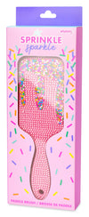 Pink rhinestone-embellished paddle brush in its retail packaging, featuring a pink handle and a clear window showing the sparkly bristles. The box has a purple header and colorful sprinkle graphics.