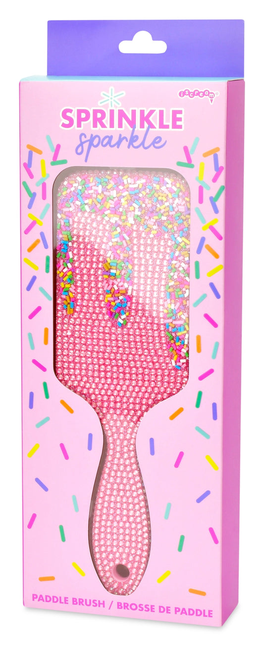 Pink rhinestone-embellished paddle brush in its retail packaging, featuring a pink handle and a clear window showing the sparkly bristles. The box has a purple header and colorful sprinkle graphics.