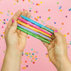 Hands holding a bundle of colorful motivational pens against a pink background with confetti dots.