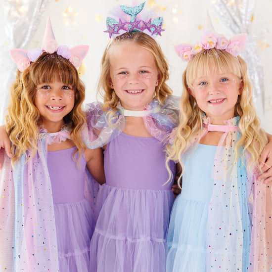 Three young girls wearing pastel unicorn and mermaid princess costume dresses, sparkly headbands, and tiara accessories, standing together against a festive silver tinsel backdrop.