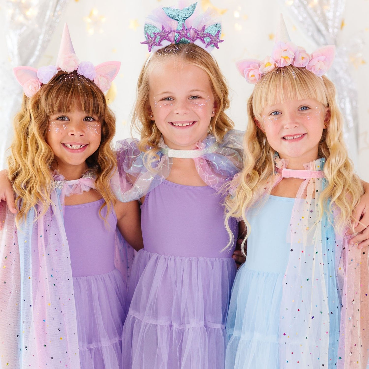 Three young girls wearing pastel unicorn and mermaid princess costume dresses, sparkly headbands, and tiara accessories, standing together against a festive silver tinsel backdrop.