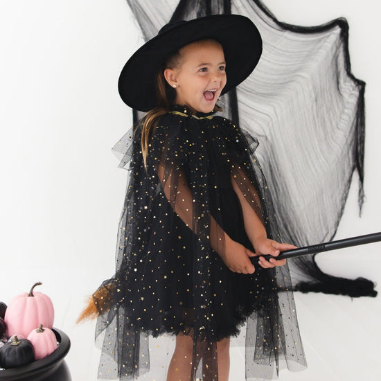 A young girl wearing a black witch costume with a wide-brimmed black hat, sheer tulle cape covered in gold glitter dots, and matching black tulle wings, holding a broomstick against a white studio background with a black pot of pumpkins at the side.
