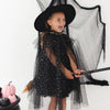 A young girl wearing a black witch costume with a wide-brimmed black hat, sheer tulle cape covered in gold glitter dots, and matching black tulle wings, holding a broomstick against a white studio background with a black pot of pumpkins at the side.