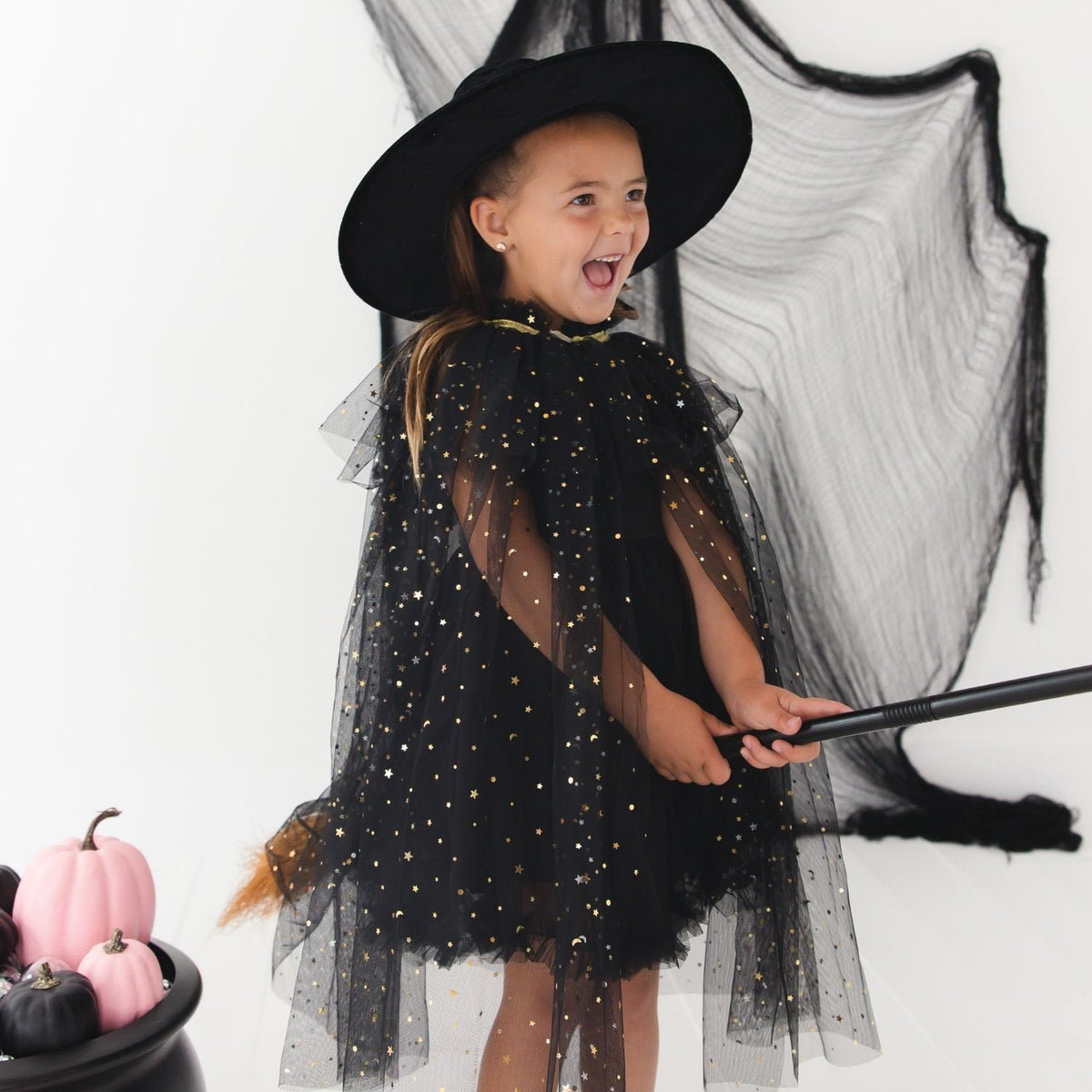 A young girl wearing a black witch costume with a wide-brimmed black hat, sheer tulle cape covered in gold glitter dots, and matching black tulle wings, holding a broomstick against a white studio background with a black pot of pumpkins at the side.