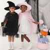 Two young girls modeling Halloween costumes, one in a black witch costume with a black hat and cape, and the other in a white dress with a pink hat and pink cape. They are standing next to a decorated pumpkin basket filled with silver foil balloons.