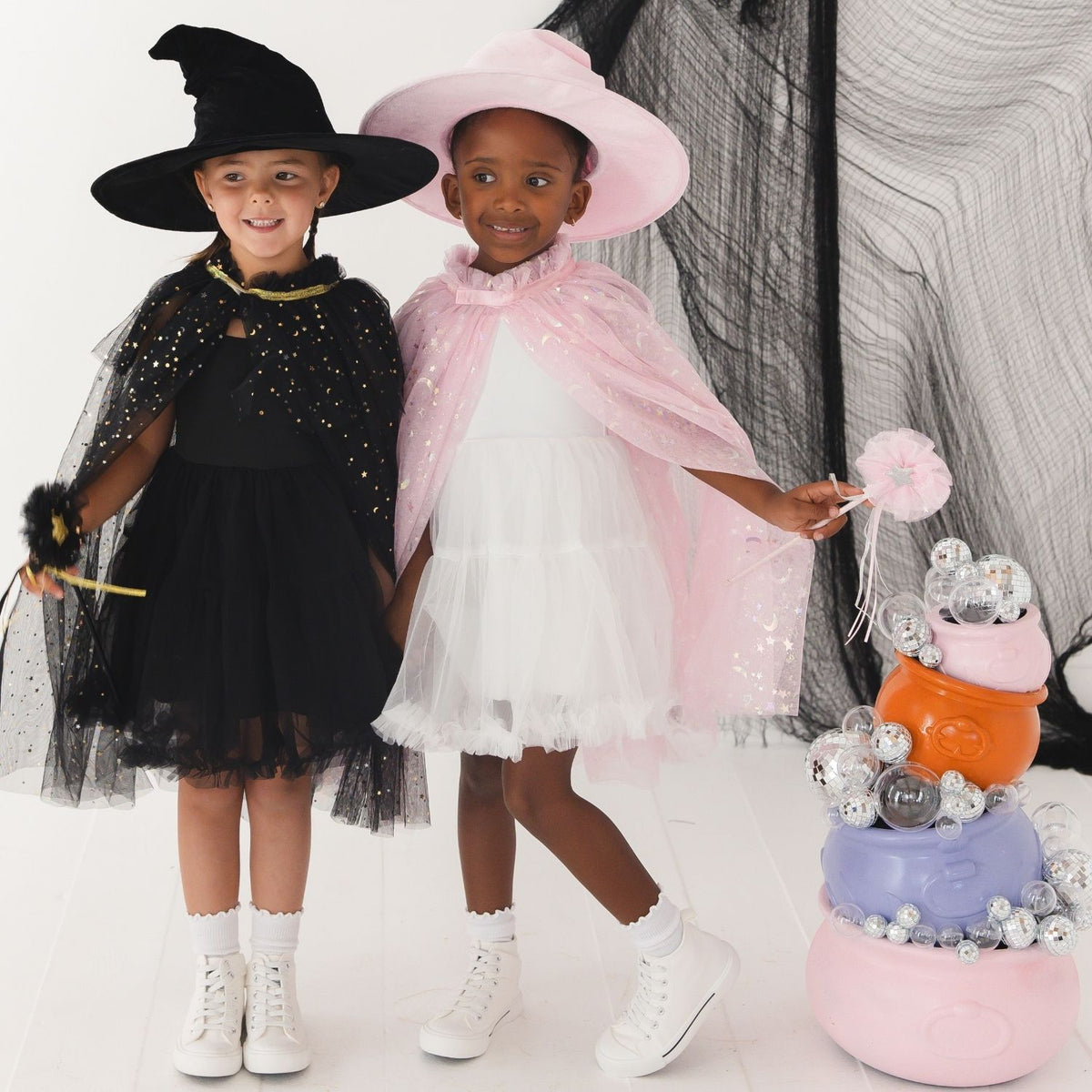 Two young girls modeling Halloween costumes, one in a black witch costume with a black hat and cape, and the other in a white dress with a pink hat and pink cape. They are standing next to a decorated pumpkin basket filled with silver foil balloons.
