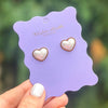 Pearl heart stud earrings displayed on a purple scalloped backer card, held in a hand with a blurred outdoor background.
