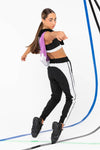 Female dancer wearing black jogger-style dance pants with white side stripes, shown mid-movement with arms raised.