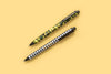 Two Taylor Elliott gel pens with camo and gingham designs on yellow background