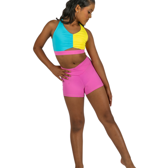 Young dancer wearing pink V-waist dance shorts with a colorful crop top, standing in a studio pose.