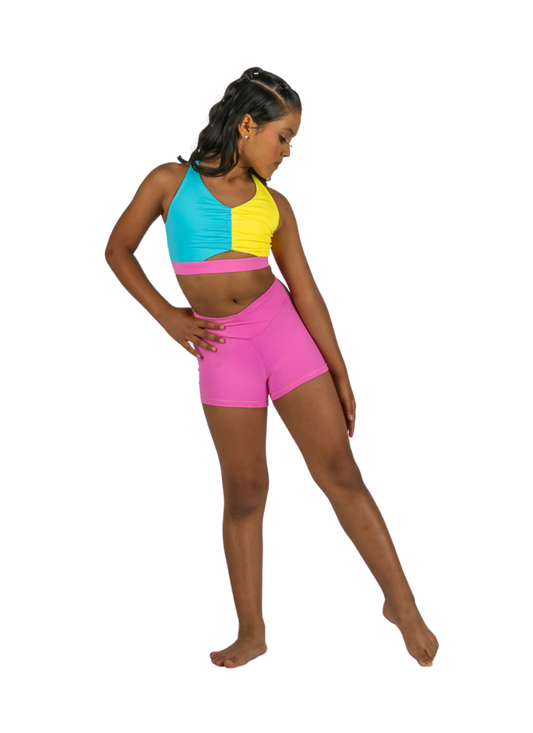Young dancer wearing pink V-waist dance shorts with a colorful crop top, standing in a studio pose.