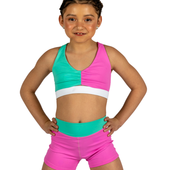 Young dancer wearing teal and pink dance shorts with matching crop top, standing facing forward.