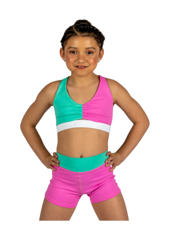 Young dancer wearing teal and pink dance shorts with matching crop top, standing facing forward.