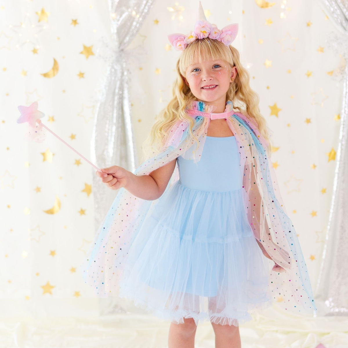 A young girl with blonde hair wearing a light blue fairy costume dress with a sheer pastel rainbow cape, a pink unicorn headband, and holding a star wand, posing in front of a white curtain decorated with gold stars.