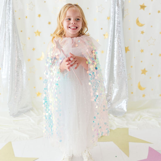 A young girl stands smiling in a sparkly white dress and holding a wand, wearing a pastel pink sequined angel cape. She is posed in front of a festive backdrop featuring silver sequin curtains, gold stars, and gold crescent moons on a white background.