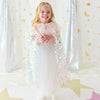 A young girl stands smiling in a sparkly white dress and holding a wand, wearing a pastel pink sequined angel cape. She is posed in front of a festive backdrop featuring silver sequin curtains, gold stars, and gold crescent moons on a white background.