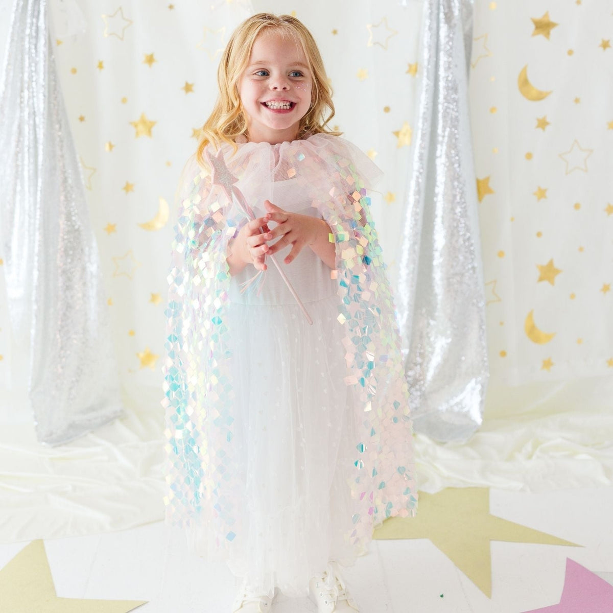 A young girl stands smiling in a sparkly white dress and holding a wand, wearing a pastel pink sequined angel cape. She is posed in front of a festive backdrop featuring silver sequin curtains, gold stars, and gold crescent moons on a white background.
