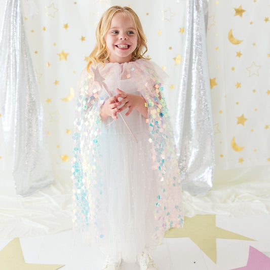 A young girl stands smiling in a sparkly white dress and holding a wand, wearing a pastel pink sequined angel cape. She is posed in front of a festive backdrop featuring silver sequin curtains, gold stars, and gold crescent moons on a white background.