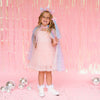 A young girl stands in a pink room wearing a pink dress, a sparkly blue and lavender cape, a matching headband with cat ears, and white sneakers. The background features a pink tinsel curtain and scattered silver disco balls.