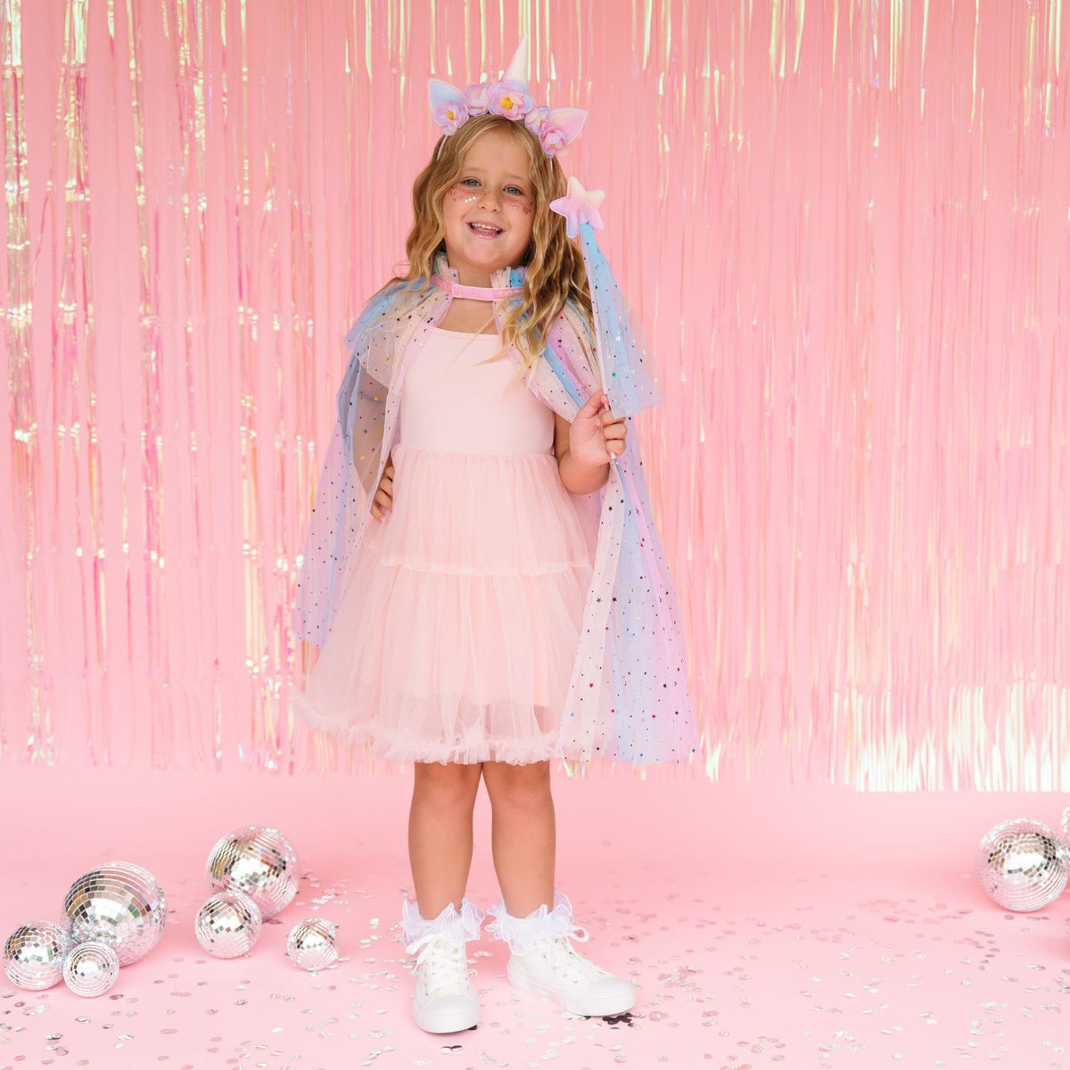 A young girl stands in a pink room wearing a pink dress, a sparkly blue and lavender cape, a matching headband with cat ears, and white sneakers. The background features a pink tinsel curtain and scattered silver disco balls.