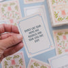 A hand holds a square white card with a light blue border and the phrase 'DANCE LIKE YOUR ARMS ARE NOODLES AND YOUR LEGS ARE STIFF.' in dark blue text.