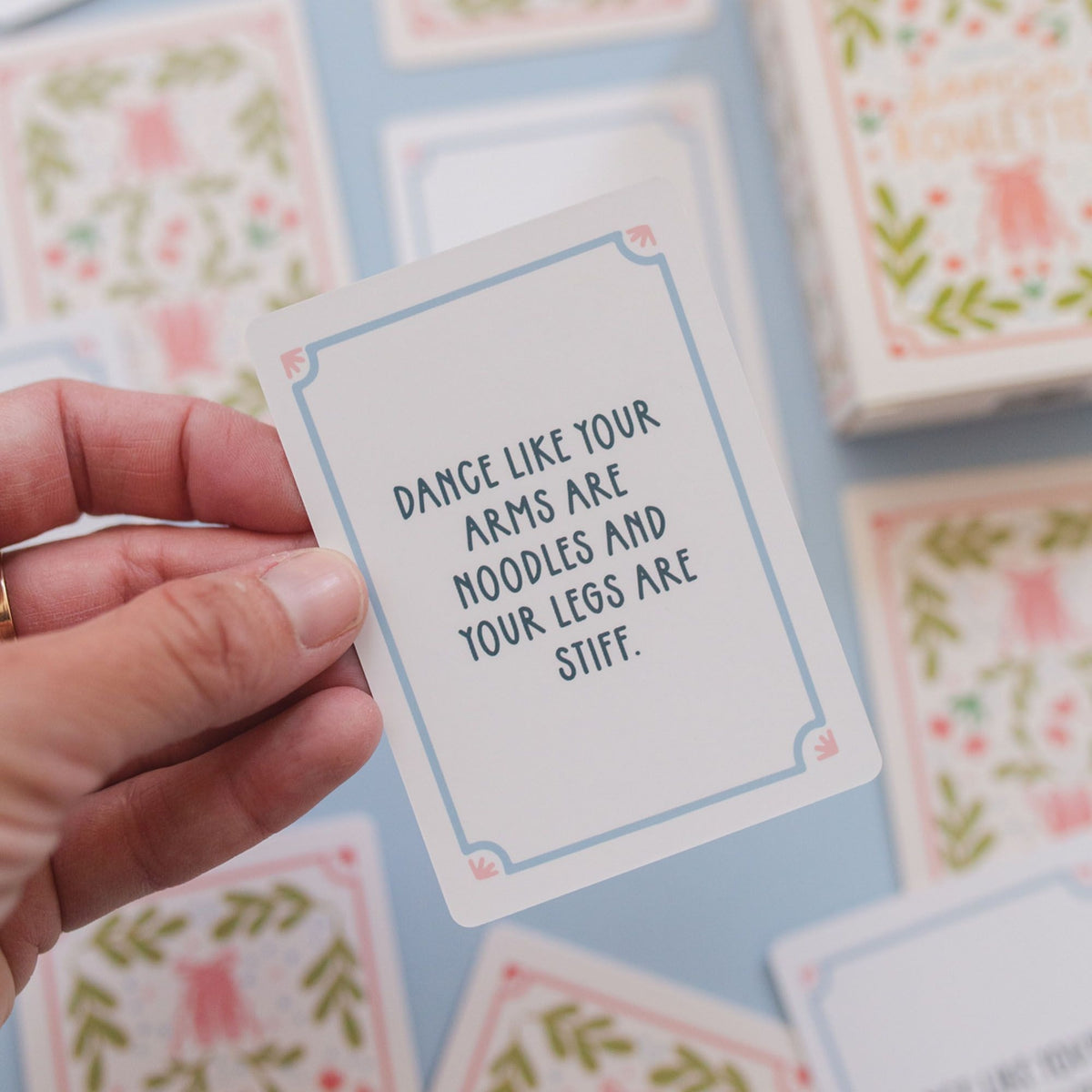 A hand holds a square white card with a light blue border and the phrase 'DANCE LIKE YOUR ARMS ARE NOODLES AND YOUR LEGS ARE STIFF.' in dark blue text.