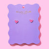 Pink heart stud earrings displayed on a purple scalloped backer card with Taylor Elliot Designs branding.