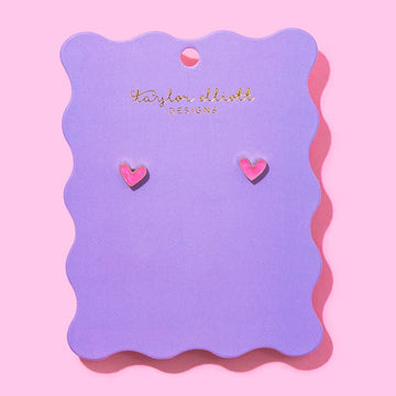Pink heart stud earrings displayed on a purple scalloped backer card with Taylor Elliot Designs branding.