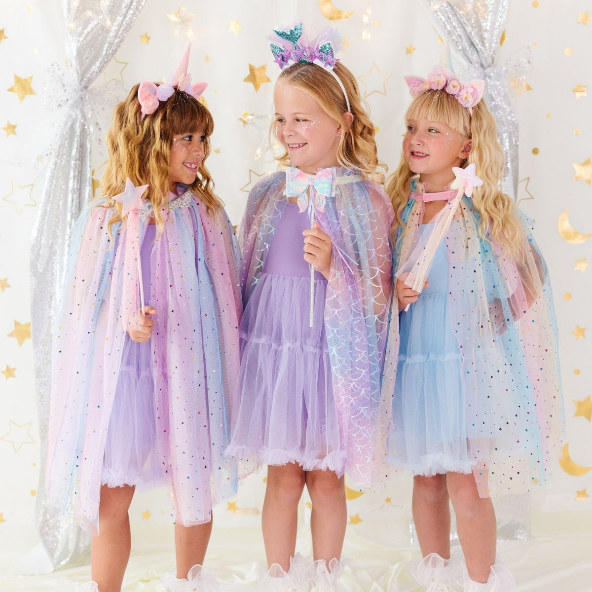 Three young girls wearing matching pastel unicorn-themed tulle capes and matching headbands, standing together in a decorated studio with silver curtains and gold star and moon decorations.