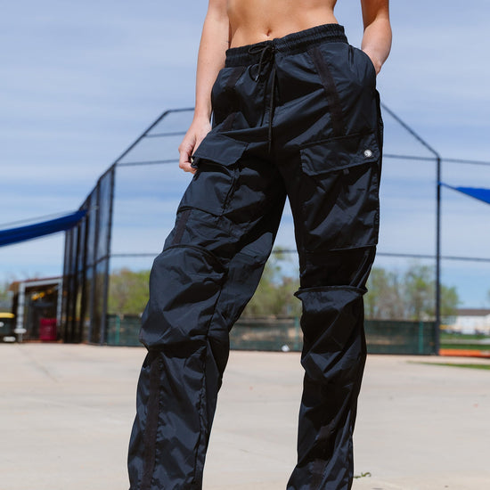 Adult dancer wearing black cargo-style dance pants, standing outdoors with hands on hips.