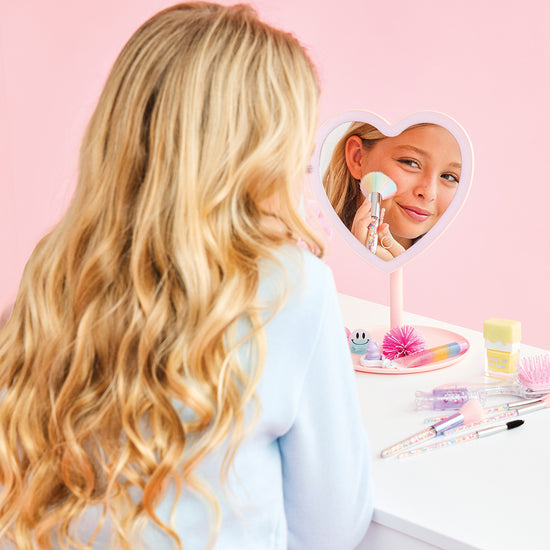 Person with long blonde hair looking into a heart-shaped mirror with a pink stand on a pink background.