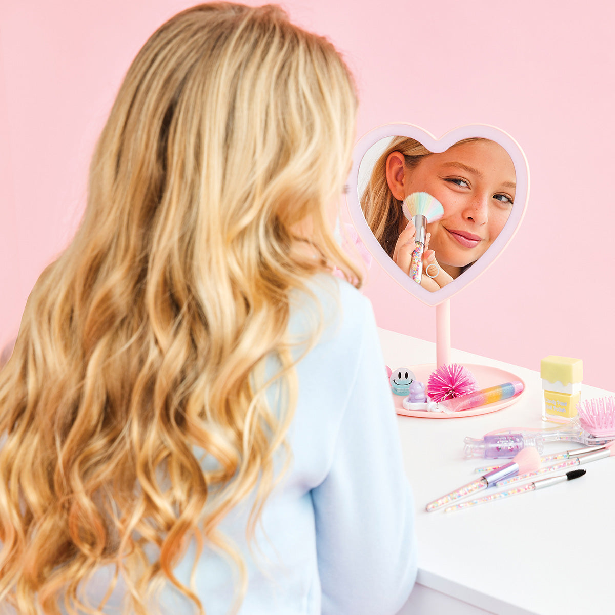 Person with long blonde hair looking into a heart-shaped mirror with a pink stand on a pink background.