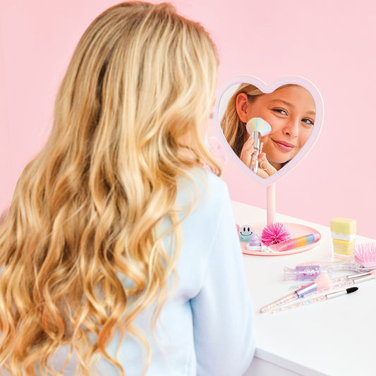 Person with long blonde hair looking into a heart-shaped mirror with a pink stand on a pink background.