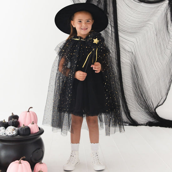 A young girl modeling a black witch costume with a sheer black tulle cape, black dress, black pointed hat, and a gold star wand, standing in a studio with a white background, black netting backdrop, and a black cauldron with pumpkins.