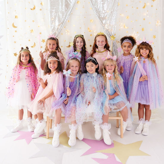 A group of young girls wearing matching pastel princess dresses and sparkly cape costumes, posed together in a decorated studio with silver curtains, gold crescent moon accents, and star-shaped floor decorations.