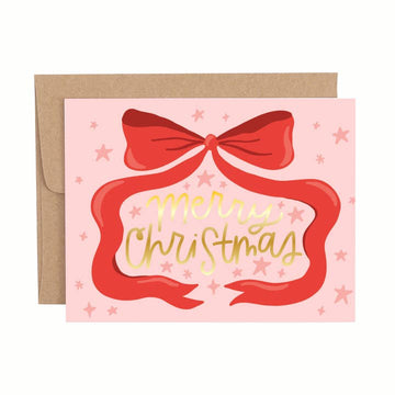 Christmas greeting card with a large red bow illustration, gold foil “Merry Christmas” lettering, and pink background with star accents, shown with a brown envelope.