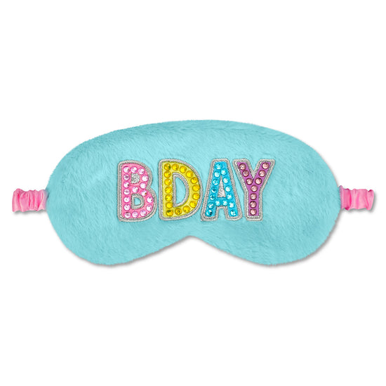 A light blue fuzzy sleep mask with elastic straps on both sides, featuring the word 'BDAY' spelled out in colorful rhinestone-embellished letters across the front, set against a plain white background.