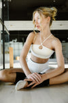 Adult dancer wearing a light-colored dance bra with matching bottoms, seated indoors.