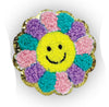 A round embroidered patch shaped like a flower with a yellow smiley face in the center and multicolored textured petals in pink, purple, and teal, outlined with a gold glittery border on a white background.
