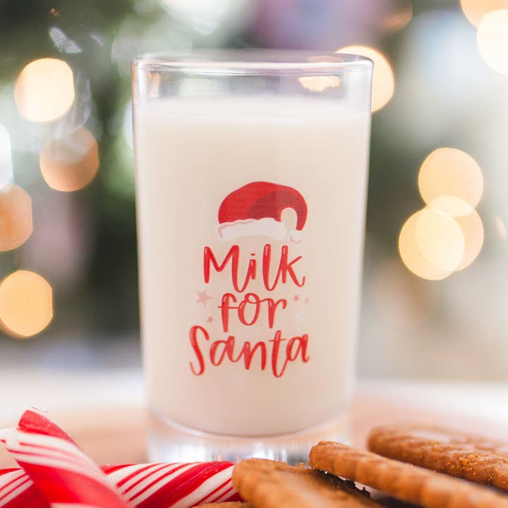 Clear glass with “Milk for Santa” design filled with milk beside Christmas cookies and lights