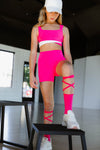 Adult dancer wearing bright pink dance shorts with a matching top, standing indoors on a stool.
