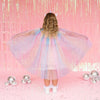 A child viewed from behind wearing a pastel iridescent tulle costume dress with a large spread skirt, standing on a pink floor in front of a pink tinsel backdrop with silver reflective balls around them.
