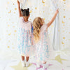 Two young girls seen from behind wearing matching iridescent sequin dresses and white sneakers, standing in front of a white curtain decorated with gold stars and crescent moons.