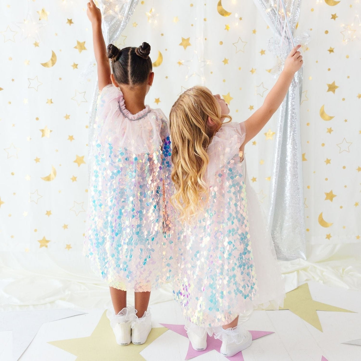 Two young girls seen from behind wearing matching iridescent sequin dresses and white sneakers, standing in front of a white curtain decorated with gold stars and crescent moons.